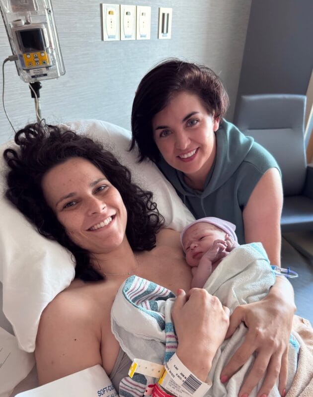 New mother with curly dark hair holding newborn Leo Alexander in a hospital bed, with a smiling woman with short dark hair leaning in beside her