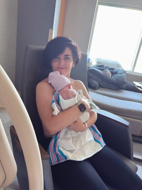Cathryn Lavery seated in a hospital room chair holding swaddled newborn Leo Alexander, who is wearing a pink striped hospital hat